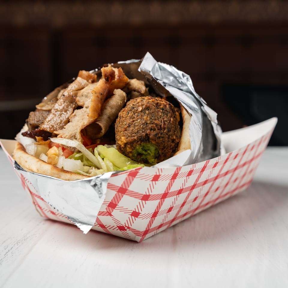 A gyro and falafel pita sandwich in a red and white checkered paper tray.