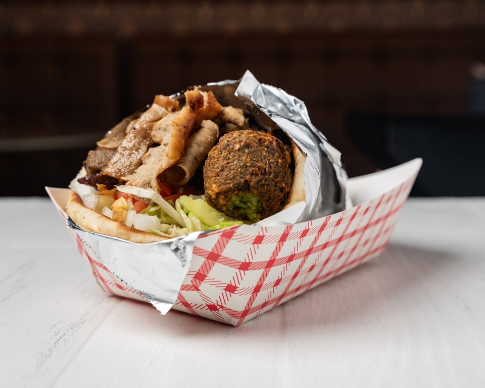 A gyro and falafel pita sandwich in a red and white checkered paper tray.