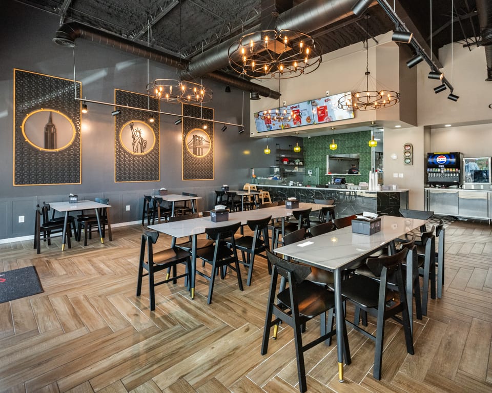 Modern restaurant interior with dark gray walls, light wood floors, black tables, and New York City themed decor.