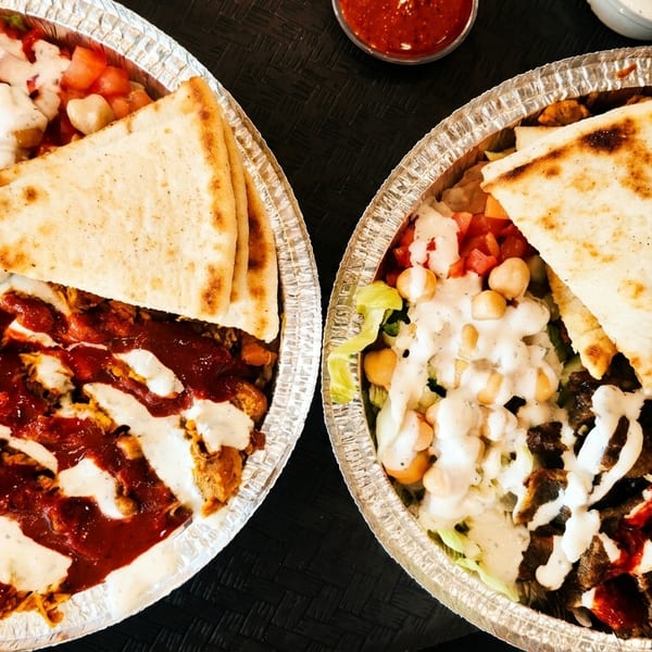 Two foil containers filled with chicken and lamb shawarma, rice, salad, pita bread, and sauces.