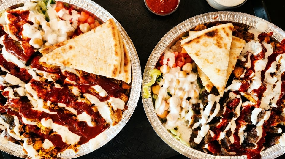 Two foil containers filled with chicken and lamb shawarma, rice, salad, pita bread, and sauces.