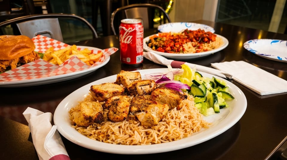 Plates of chicken and rice, a burger with fries, and a soda on a dark table.